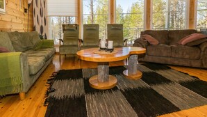 Een gezellig chaletinterieur met houten wanden, een ronde houten tafel, twee groene fauteuils, een bruine bank en grote ramen met uitzicht op bomen.