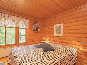 Un dormitorio de cabaña de madera con una cama, una ventana, una estantería con libros y un cuadro en la pared.