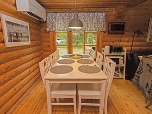 A wooden cabin interior with a dining table, chairs, a hanging light fixture, and a view through the window.