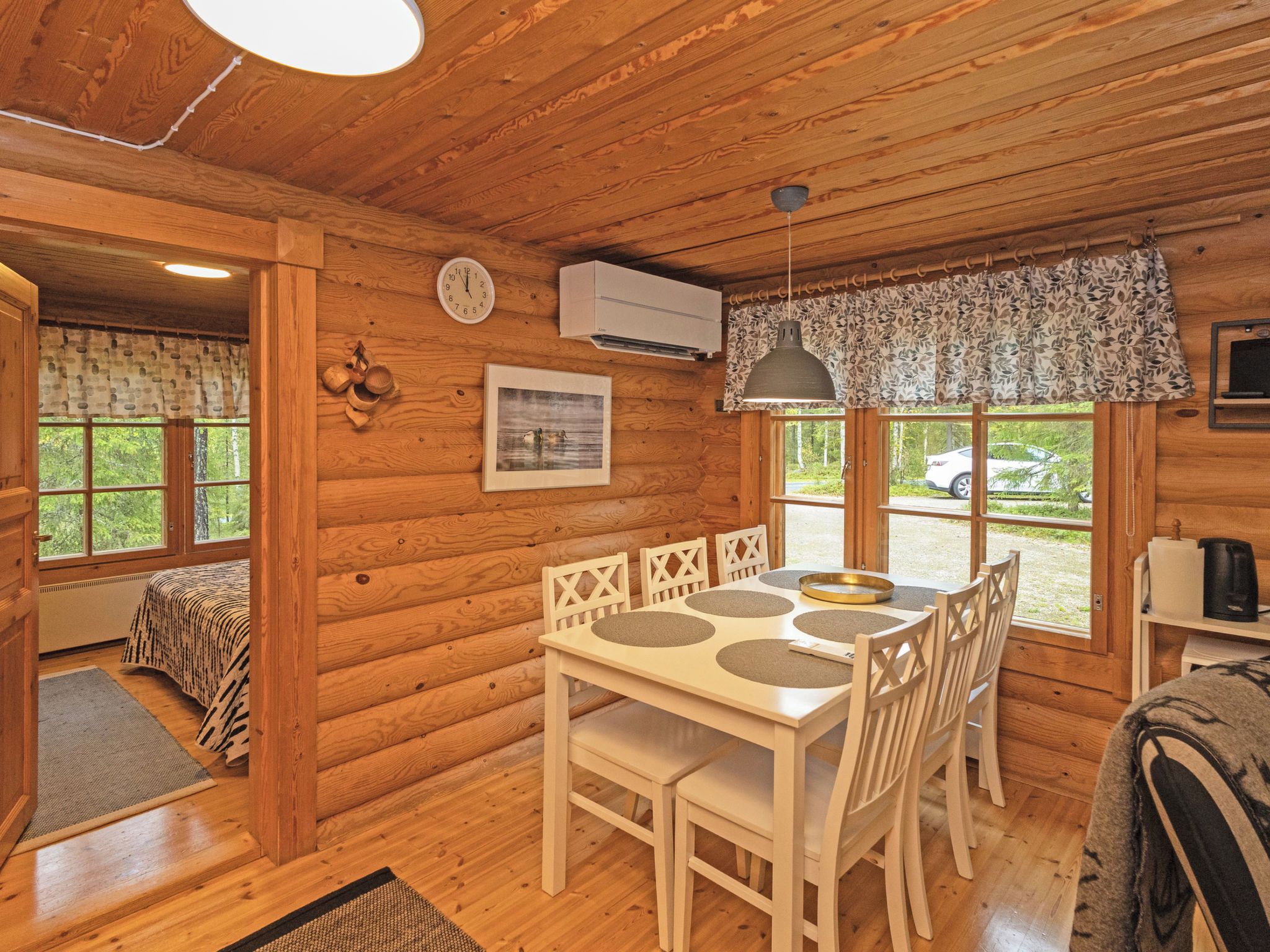 Interior de una cabaña de madera con una mesa de comedor, sillas, un reloj, un equipo de aire acondicionado y una ventana con vistas al exterior.