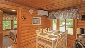 Interior de una cabaña de madera con una mesa de comedor, sillas, un reloj, un equipo de aire acondicionado y una ventana con vistas al exterior.