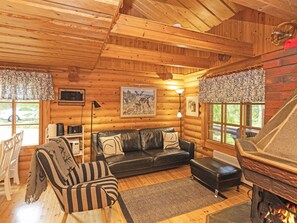 A cozy cabin interior with a wooden ceiling, a black leather sofa, a striped armchair, a fireplace, and a dining area with a table and chairs.