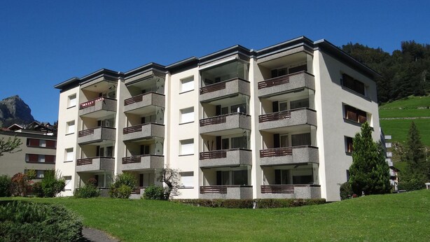 Un edificio residencial de varios pisos con balcones, ubicado en una ladera con cielo despejado.