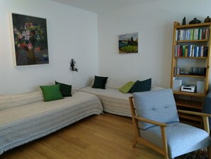 Ein Wohnzimmer mit einer Sofagarnitur, einem Sessel, einem Bücherregal und zwei Bildern an der Wand.