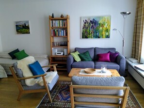 A living room with a sofa, armchair, coffee table, bookshelf, and a painting on the wall.