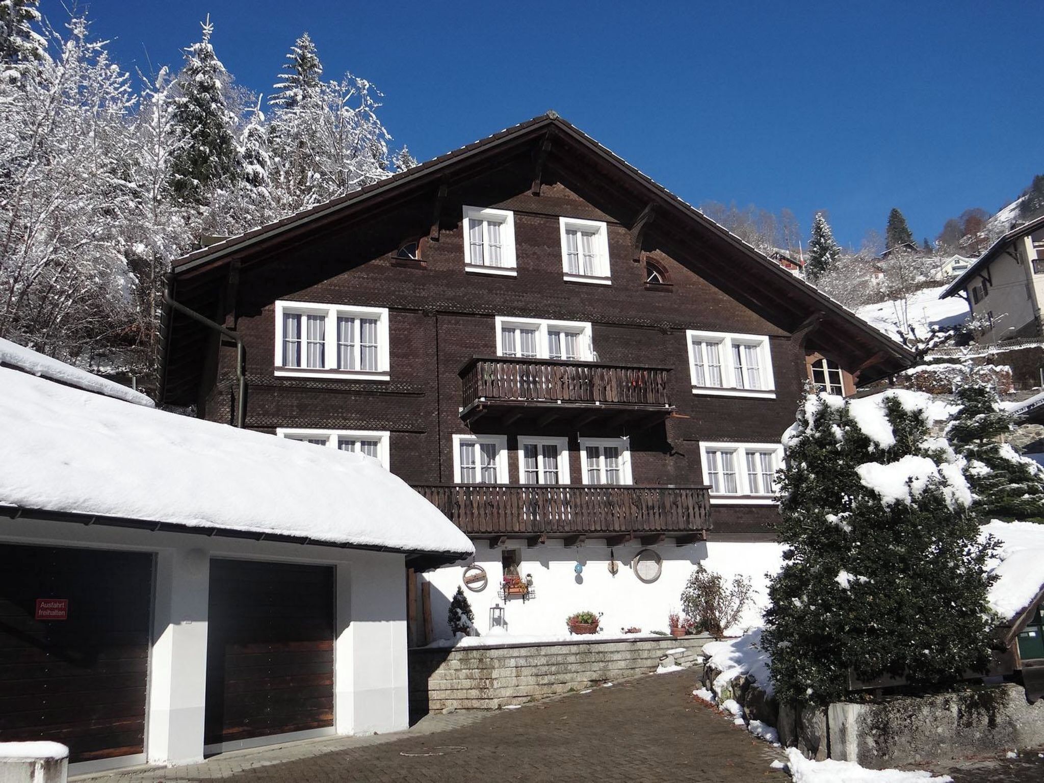 A traditional chalet-style building with multiple windows and a balcony, surrounded by snow-covered trees.