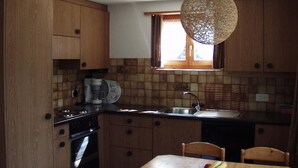 A kitchen with wooden cabinets, a built-in oven, a coffee maker, and a hanging light fixture. - Teleboy by Interhome (Lenk)