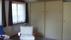 A bedroom with a bed, a chair, a wardrobe, and a window with curtains. - Teleboy by Interhome (Lenk)
