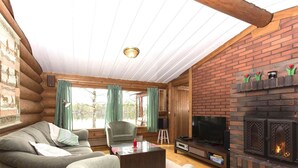 L'intérieur d'une cabane en rondins, avec un canapé, un fauteuil, un téléviseur, une cheminée et une vue sur un lac, visible à travers de grandes fenêtres.