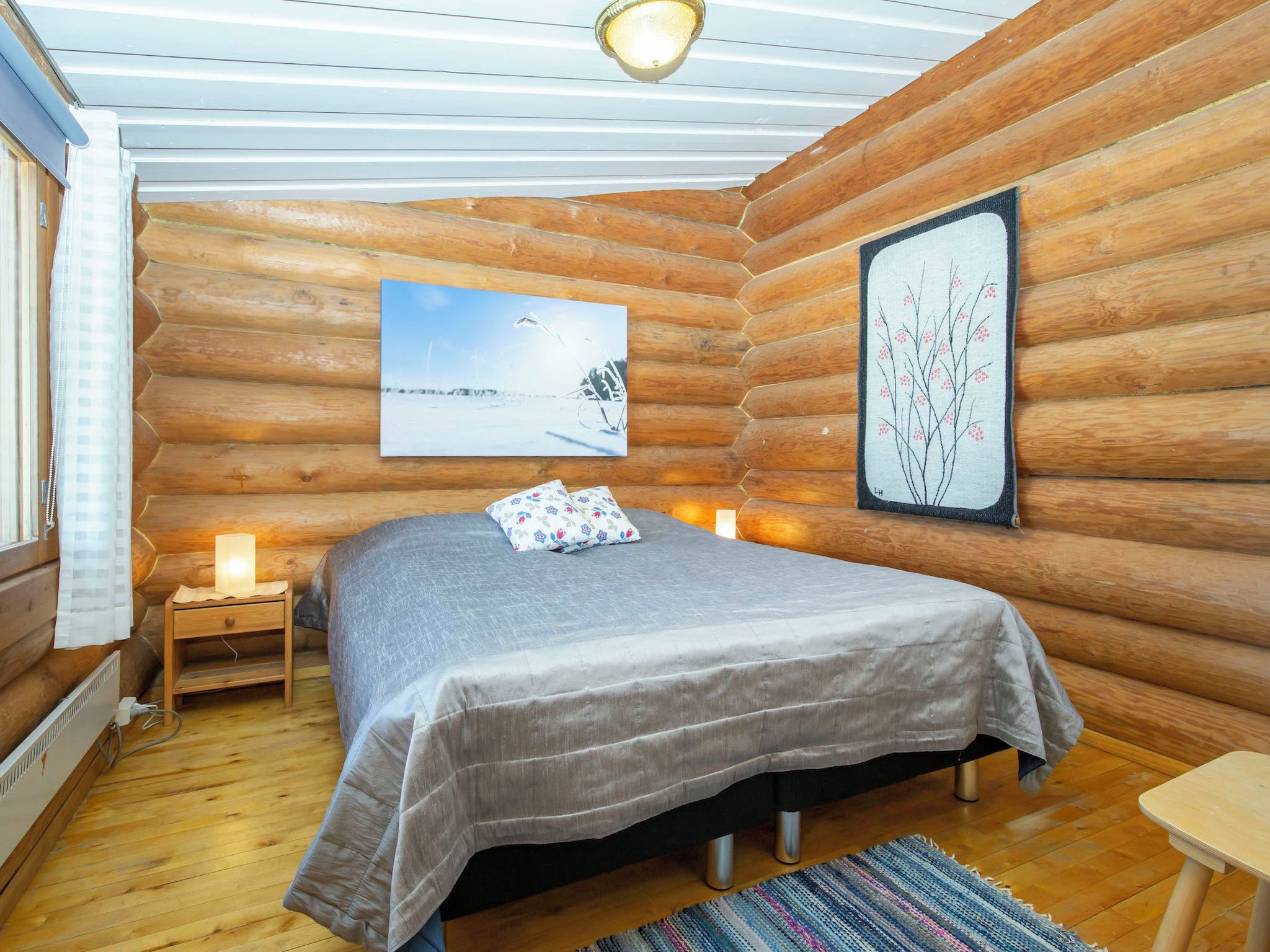 Une chambre à coucher dans un chalet en rondins, avec un lit, des lampes de chevet, un tableau encadré et une fenêtre avec des rideaux.