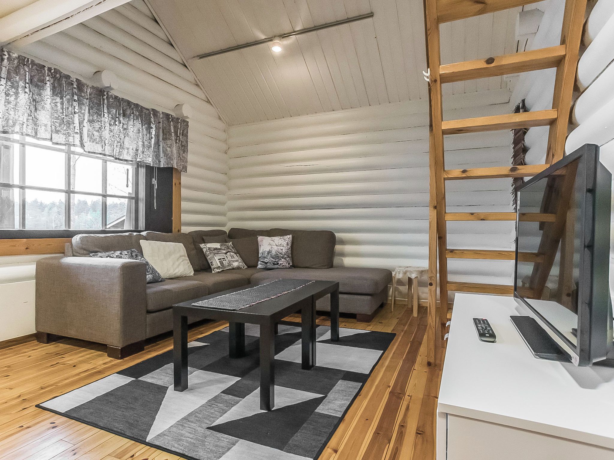 Interior de uma cabana de troncos com sofá, mesa de centro, televisão e escada de madeira.