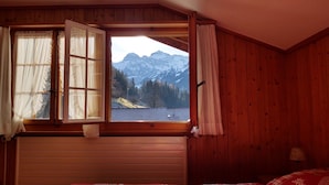 A bedroom with a large window offering a mountain view, wooden walls, and a bed with a checkered bedspread. - Bärnermutz # 2 by Interhome (Lenk)