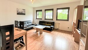 Un salon moderne avec un canapé en cuir noir, une table basse en bois et un téléviseur posé sur un meuble. Trois fenêtres laissent entrer la lumière naturelle et offrent une vue sur la verdure environnante.