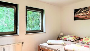 Une chambre avec deux fenêtres, un lit agrémenté d’oreillers à motifs floraux, une table de chevet avec une lampe et une affiche accrochée au mur.