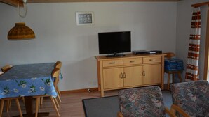 Ein Wohnzimmer mit Esstisch, zwei gemusterten Sesseln, einer Holzkommode mit Flachbildfernseher und einer Pendelleuchte.