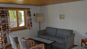 Ein gemütliches Zimmer mit einer Couch, zwei Stühlen, einem kleinen Tisch, einer Lampe und einem fenster mit Vorhängen.