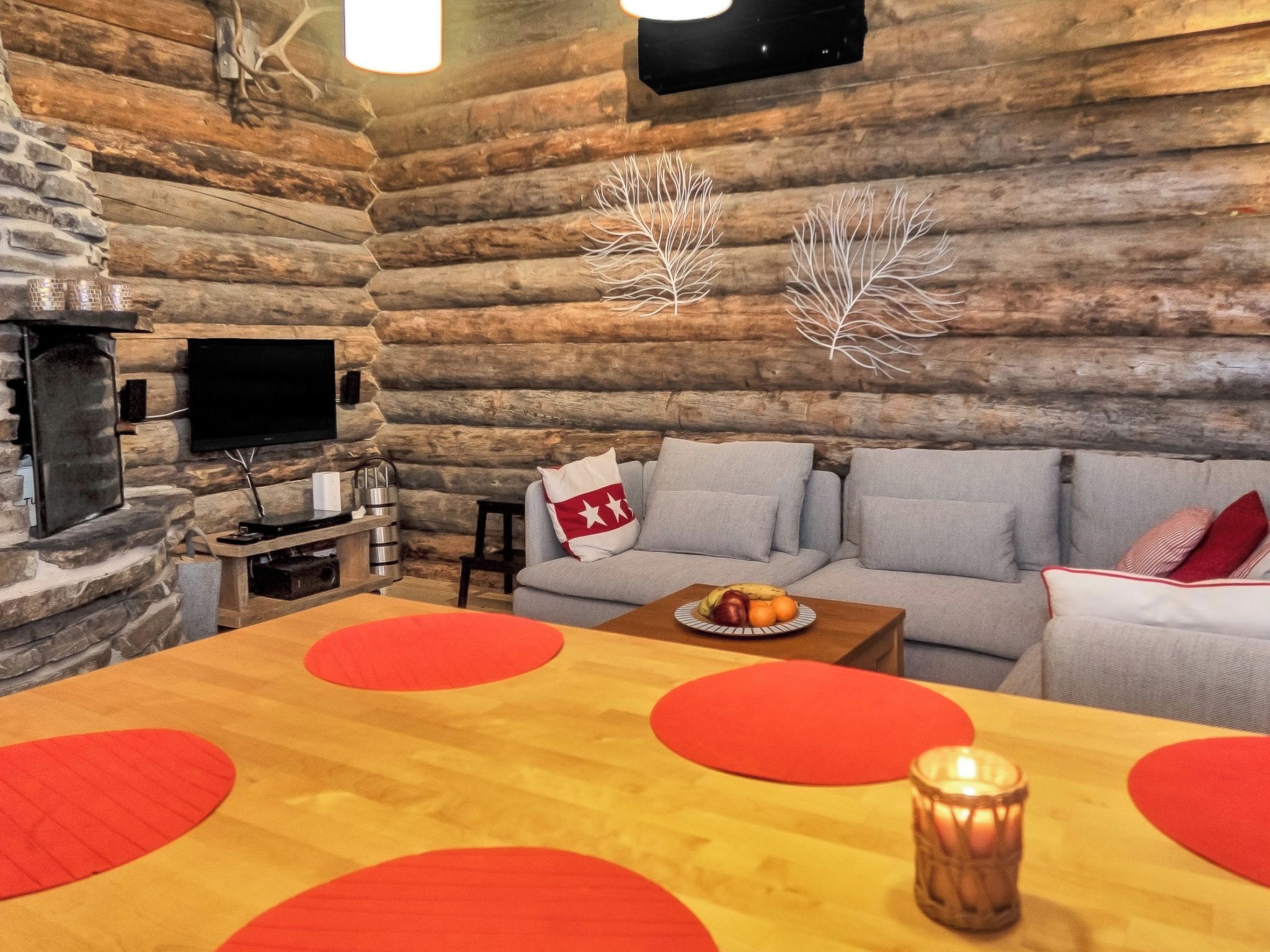 Ein Holzblockhaus-Interieur mit Kamin aus Stein, einem an der Wand befestigten Fernseher, einer Sofagarnitur und einem Esstisch mit roten Kreismustern.