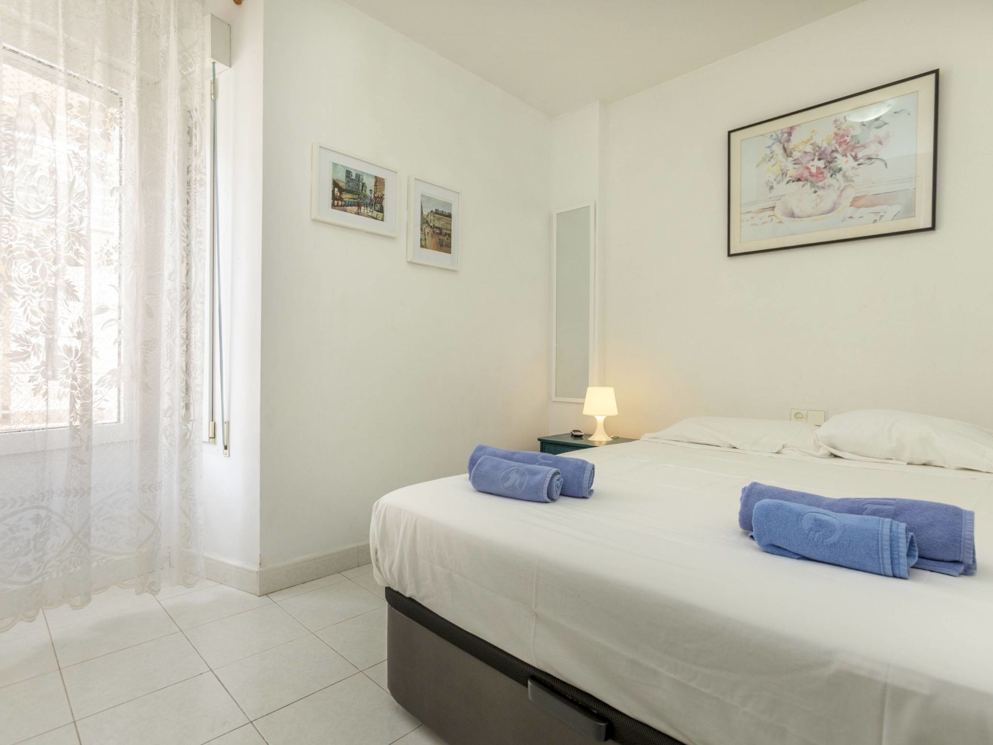 Una cama bien hecha con dos toallas dobladas, una lámpara de noche y dos cuadros en la pared.