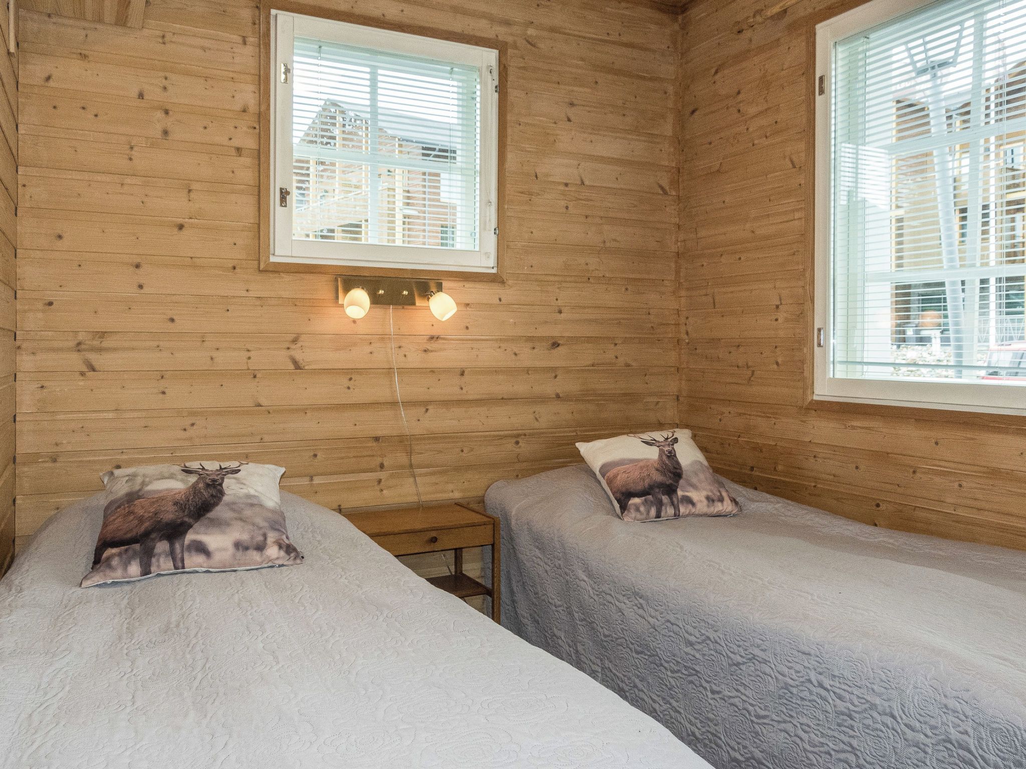 Puinen mökkihuone, jossa on kaksi sänkyä. Jokaisessa sängyssä on pehmustettu tyyny, jossa on kuvioitu peura. Huoneessa on kaksi ikkunaa, joissa on sälekaihtimet, ja sänkyjen yläpuolella seinään kiinnitetty valaisin.