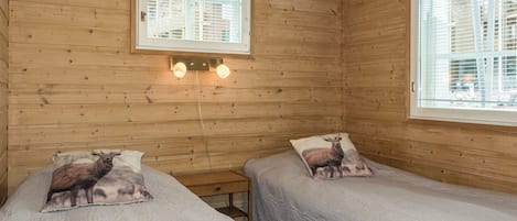 A wooden cabin room with two beds, each with a pillow featuring a deer. There are two windows with blinds, and a wall-mounted light fixture above the beds.