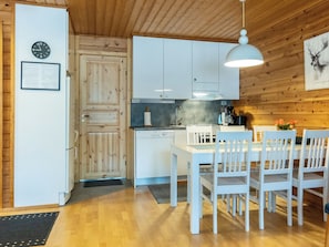 Une cuisine avec des meubles en bois, une table à manger blanche avec des chaises, un luminaire suspendu et une horloge au mur.