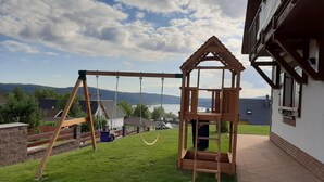 Un patio trasero con un columpio, una casita de juegos de madera y vistas a casas y montañas.