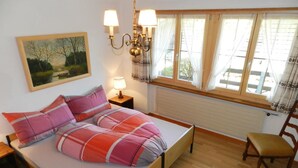 A bedroom with a bed, a nightstand, a chair, a window with curtains, a wall-mounted lamp, and a painting. - Apartment Bolgen by Interhome (Zweisimmen)