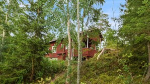 Une cabane rouge nichée dans une zone boisée, dotée d’une terrasse visible et d’un escalier menant à l’entrée.