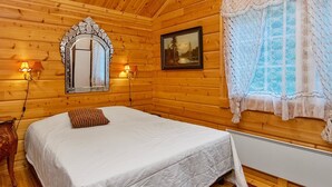 Une pièce de style cabane en bois, avec un lit, deux lampes fixées au mur, un miroir, un tableau encadré et une fenêtre ornée de rideaux en dentelle.