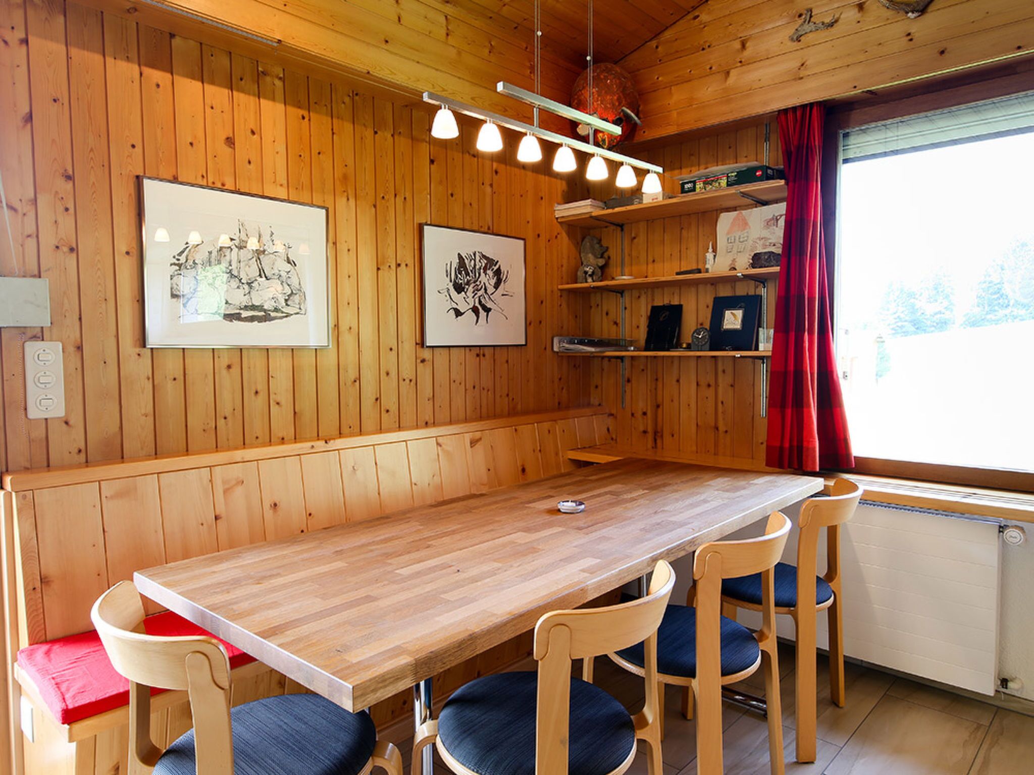 Un intérieur en bois avec une longue table et quatre chaises, des œuvres d’art encadrées accrochées aux murs et une fenêtre ornée d’un rideau rouge.