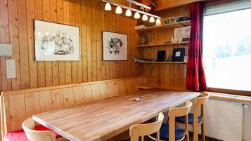 Un intérieur en bois avec une longue table et quatre chaises, des œuvres d’art encadrées accrochées aux murs et une fenêtre ornée d’un rideau rouge.