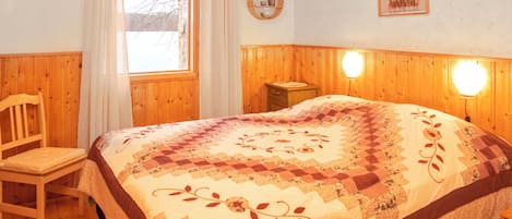 A cozy bedroom with a wooden headboard, a quilted bedspread, a bedside table, and a window with curtains.