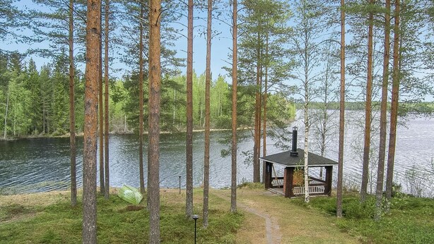 Mökki järven rannalla, jota ympäröivät korkeat puut.