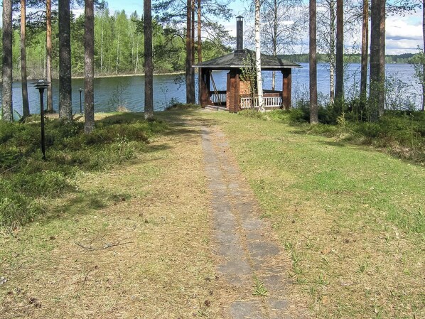 Un sentier mène à une petite cabane dotée d’une cheminée, entourée d’arbres et bordée d’un lac.