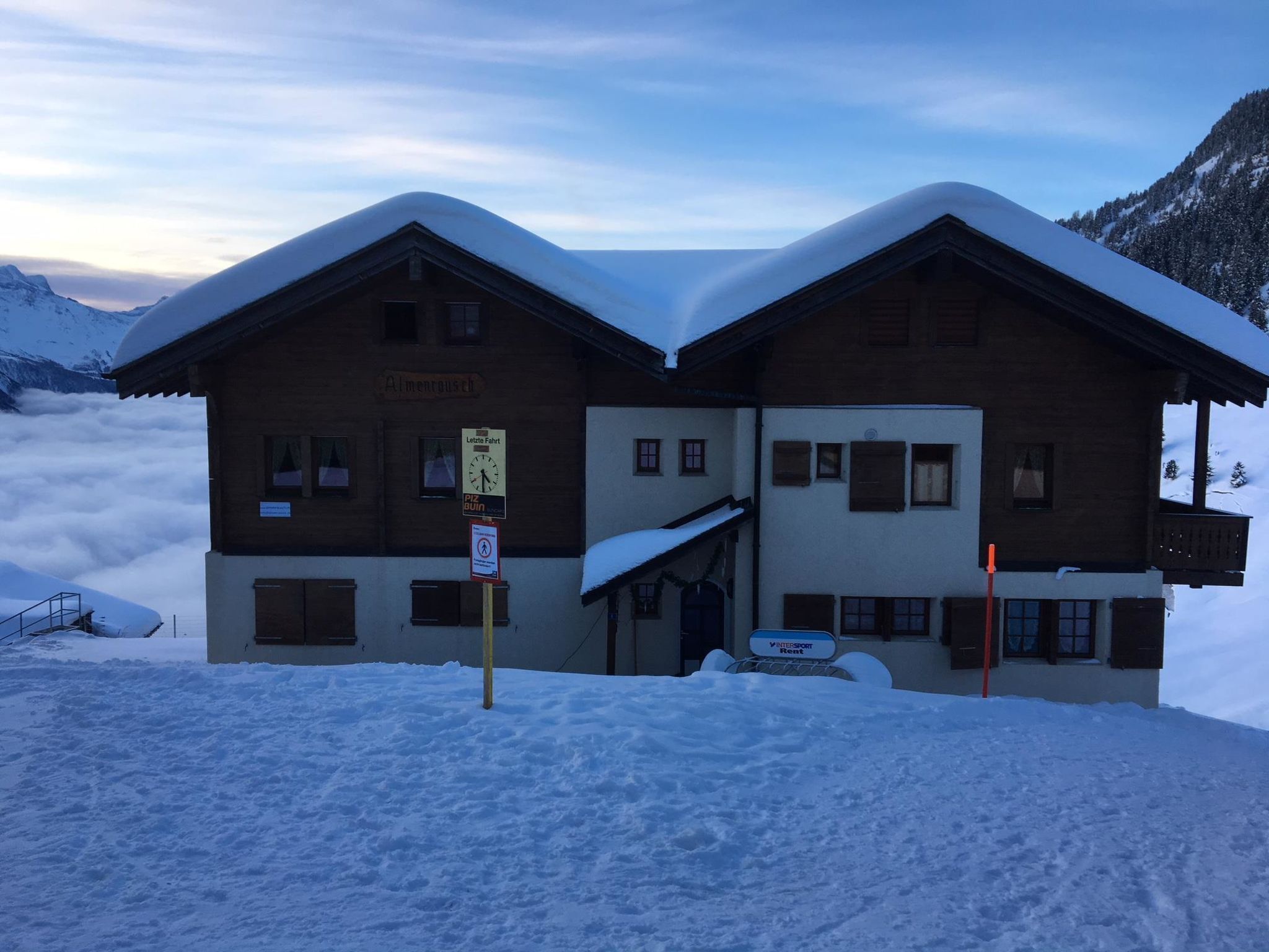 Un edificio de madera de dos pisos con techo cubierto de nieve, un letrero que indica restaurante y un cartel de alquiler de esquíes.