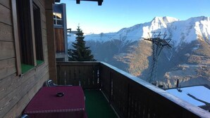 A balcony with a wooden railing, a table with a red tablecloth, and a chair. A mountain view is visible in the background.