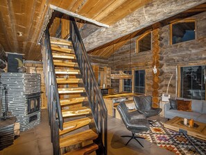 A log cabin interior with a wooden staircase, a stone fireplace, a dining area, and a cozy seating space.