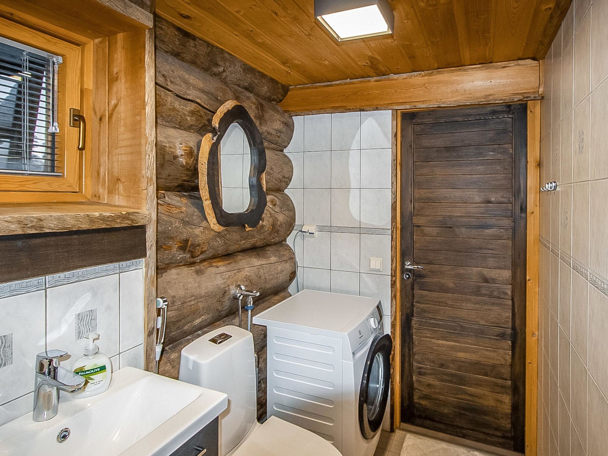 A rustic bathroom with a wooden log wall, a large mirror, a sink, a washing machine, and a door.