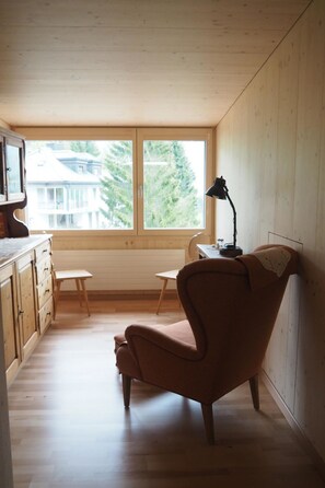 A room with a wooden floor, a chair, a desk, a lamp, and a window with a view of trees and houses. - Aurora Appartement/Fewo 4-Bettwohnung by Interhome (Engelberg)