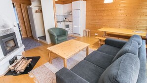 A cozy living room with a fireplace, a sofa, a wooden table, and a kitchen area with cabinets and a refrigerator. - Tunturinvieri k3 by Interhome (Ylläsjärvi)