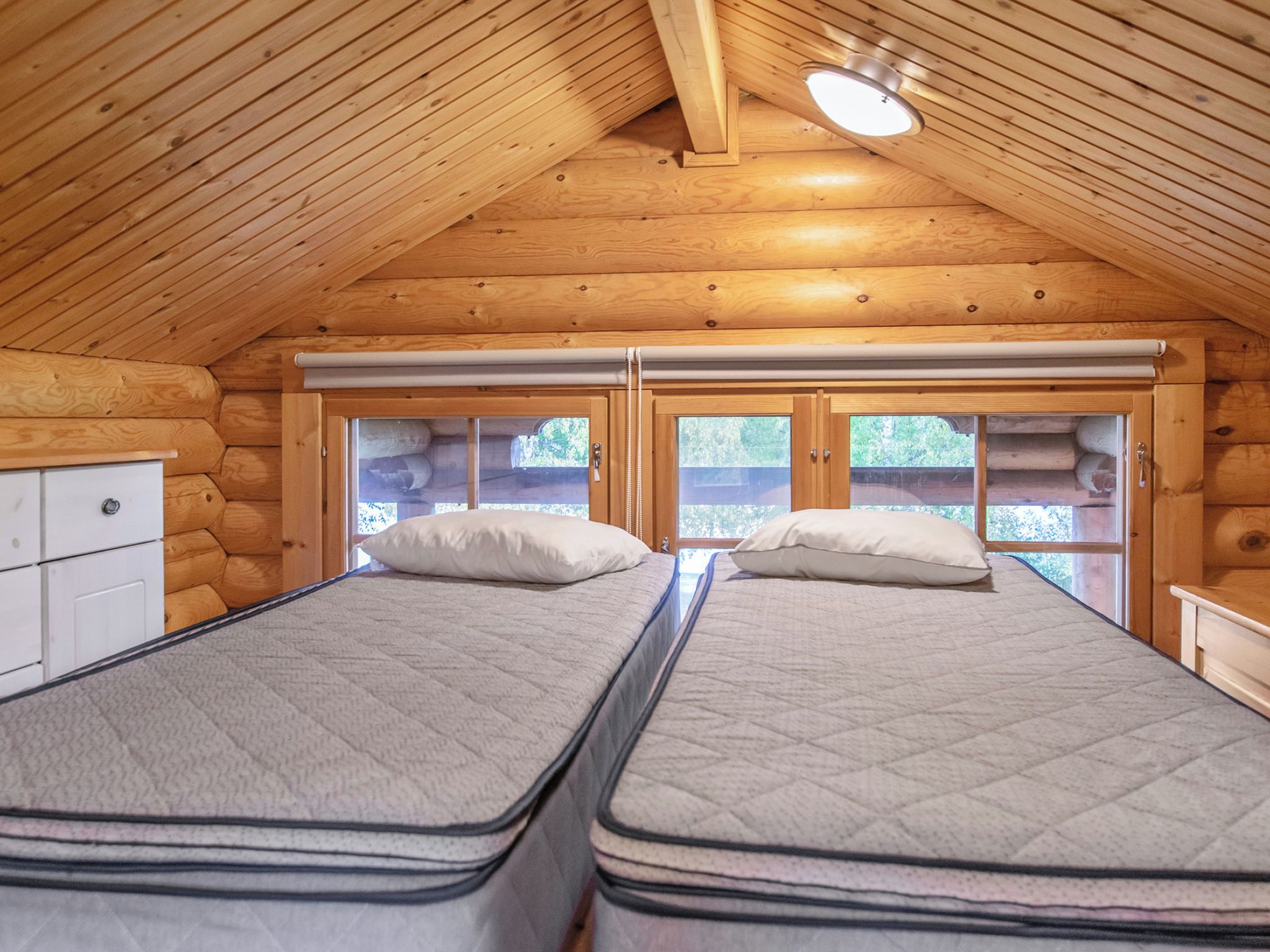 Une chambre dans un chalet en rondins, avec deux lits, une fenêtre et un luminaire au plafond.