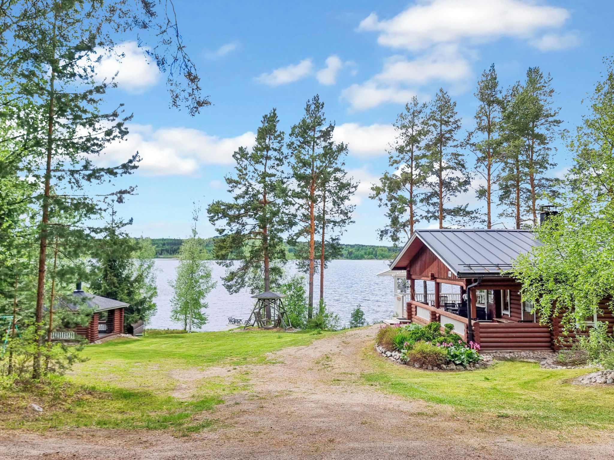 Mökki, jossa on kuisti, jolta avautuu näkymä järvelle, ja jonka ympärillä on puita.