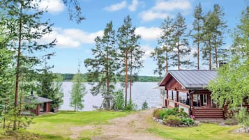 Mökki, jossa on kuisti, jolta avautuu näkymä järvelle, ja jonka ympärillä on puita.