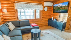Interior acogedor de una cabaña de madera con un sofá, una mesita redonda, un televisor montado en la pared y una ventana con vistas a un paisaje montañoso.