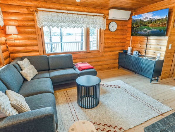 Interior acogedor de una cabaña de madera con un sofá, una mesita redonda, un televisor montado en la pared y una ventana con vistas a un paisaje montañoso.