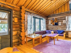 A log cabin interior with a wooden ceiling, a sofa set, a table, and a window with curtains.