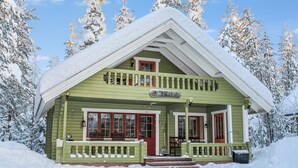 Una cabaña verde con puerta roja y balcón, rodeada de árboles cubiertos de nieve.