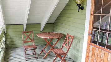 A covered porch with a wooden table and two chairs, a green exterior wall, and a window with a wooden frame.