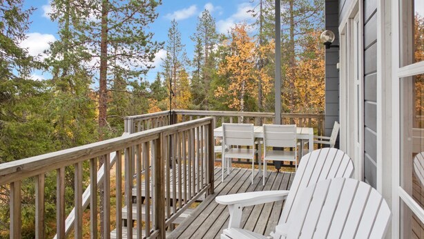 A wooden deck with white Adirondack chairs, a table, and a railing overlooking a forested area. - Vuosseli-kelkka 15a by Interhome (Kuusamo)