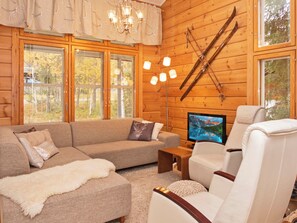 Interior de una cabaña de troncos con sofá, sillón y televisión mostrando un paisaje de montaña.